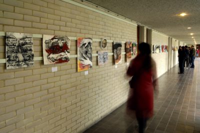 Douglas Atfield A woman in a red coat studies a row of drawings on a pale brick wall as she walks past. Her image is blurred as it is a time lapse photograph by Douglas Atfield