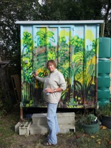Artist Ruth Hartley stands at the door of her studio made from a metal container on stilts. The front of the container is a brightly painted landscape of green and yellow plants against a light blue sky.
