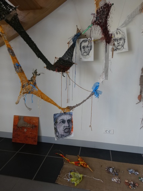 Art by Ruth Hartley: View of the Surviving Monsters installation at the Corpus exhibition at La Peleyre Gallery, France, consisting of suspended knitted scarves in brown and yellow representing the Road, black-and-white drawings hanging from the Road and shells, stones and other objects collected along the journey lying on a piece of hessian on the floor