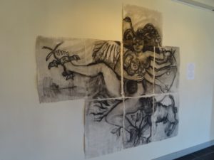 Art by Ruth Hartley: A charcoal painting of Medusa over a grid of 7 canvases. She has multiple arms, one wing, multiple breasts and an open womb. Her right leg is tied down and her left leg rooted in the ground. A powerfull looking dog is barking at her side.