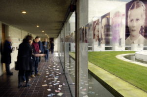 Art by Ruth Hartley: Ruth participated the Mother Art exhibition. This photo by Douglas Atfield shows visitors to the exhibition looking at postcards scattered on the floor of a long corridor alongside a glass wall with portraits hanging. The portrait nearest to the viewer is of a young girl.