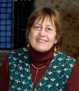 Ruth Hartley smiles at the camera. Her expression is friendly, pleased and engaging, inviting contact.She wears light-rimmed spectacles, a red-brown ribbed roll-neck top with a dark green-and-cream patterned pinafore over it, as well as ochre earrings and a black-and-ochre necklace.
