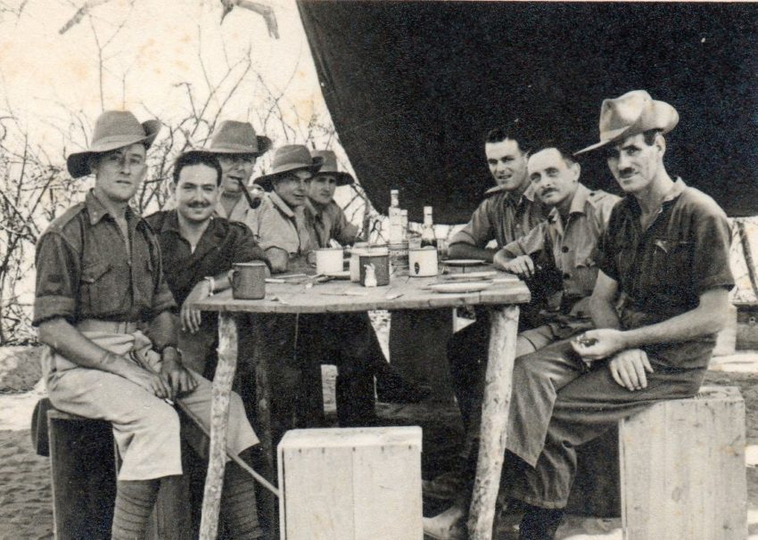 The black and white photo shows 8 men in uniform uaround a rough table under a canvas shelter - They have long shorts and felt bush hats