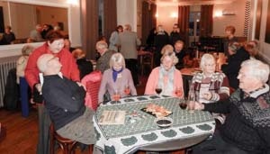 A group of people sittting around a table chatting. In the background groups of people standing and talking