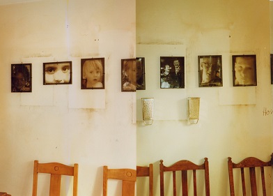 A row of chairs sit below a row of transparent family photos lined up on a wall marked by the stains made by the removal of previous photos. The eyes of all the people in the photos are lined up so that one can see the family resemblances between the children and the photos of their parents as children