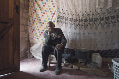 An old white-haired Zambian man in a suit with a medal bar on hs chest sits on a stool in his concrete block house with his possessions arounf him. Behind him his bed is hidden by curtains.