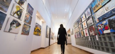 Image from the stART Foundation website A woman in black is silhouetted as she walks down a long gallery with paintings on either side