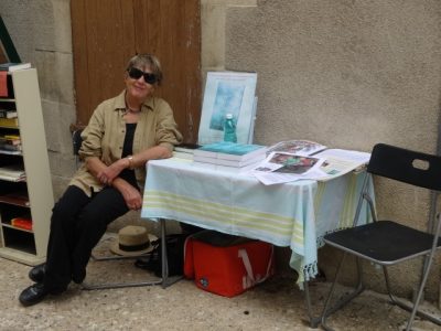 Ruth sits a table on which are her novels against a wall in the street outside a bookshop