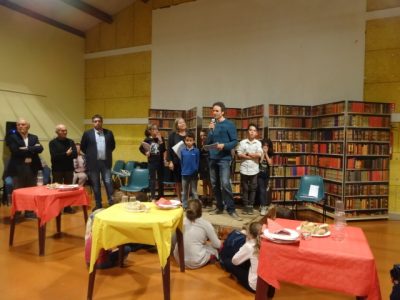 The author stands in the middle of a group of children in front of a screen that looks like a bookcase. The village mayor stands on his right.