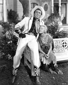 The photo shows a handsome cowboy in a snazzy white suit and stetson leaning on a garden bench on which sits a scruffy and dirty hillbilly girl with a blond plait and a dirty face. She is in love with the cowboy.