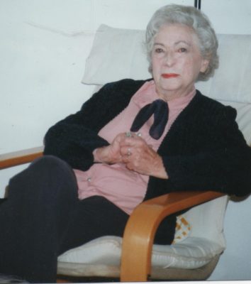 Rica sits in a Swedish-style chair wearing a black suit with a pink blouse. Her hands are clasped and her hair is white. Her face shows that she has had treatment for skin cancer.