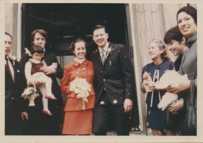 Wedding photo at the registry office door. The men are in dark suits, Ruth Hartley in a red suit, Rica Hodgson in blue, Noppy and Jean and Ruth carry bouquets, Rachma has a basket of flowers and a brown pinafore.
