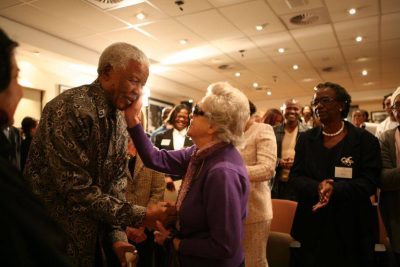 Rica Hodgson affectionately cups the cheek of Nelson Mandela with her right hand as he leans toward her, grasping her left hand with his right. The are at a function where several people in the background appear delighted with this display of affection.