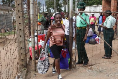 A young woman grinning happily walks out of the prison gates at Chikarubi watched by a prison officer