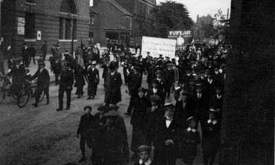 A group of working-class men, women and children in their best clothes walk down Bow Road with placards and banners in the rear.