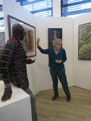 Cynthis zukas gestures at a painting in the Lechwe Trust Gallery. Wiulliam Miko is on the left