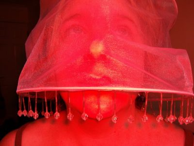 Red light suffused a phtograph of a woman's face obscured by a red transparent headdress with beads on it