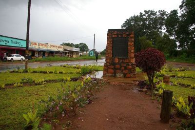 The photo shows the memeorial in a lsightly scruffy park after a tropical rainstorm. In the background are single story African shops with verandahs typical of a small African town