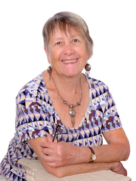 Ruth Hartley smiles at the camera. Her expression is friendly, pleased and engaging, inviting contact. She wears light-rimmed spectacles, a red-brown ribbed roll-neck top with a dark green-and-cream patterned pinafore over it, as well as ochre earrings and a black-and-ochre necklace.