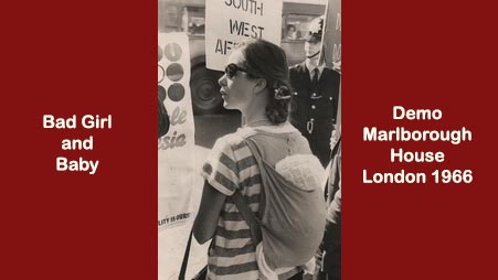 Black and white image of a young woman in a striped t-shirt with baby in a cloth carrier on her back at at demonstration outside Marlborough House in London in 1966. She wears sunglasses and had her hair tied back. A policeman stands in the background next to a placard bearing the words SOUTH WEST AF...