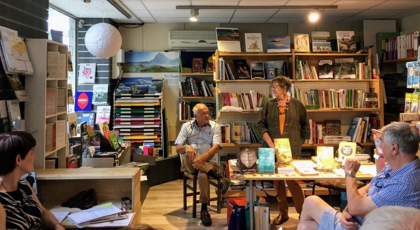 Before the start of the launch of the books that took a lifetime to write, John Corley the MC is seated behind the book table while author Ruth Hartley, standing, turns toward him.