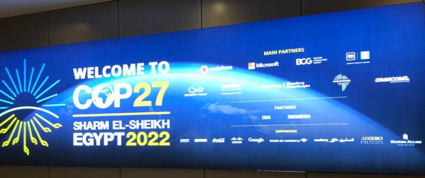 A huge blue billboard with a white, blue and yellow sunburst logo and white and yellow text says WELCOME TO COP27, SHARM EL-SHEIKH, EGYPT2022, with the logos of 20 partners including Microsoft and Google displayed to the right of the welcome message.