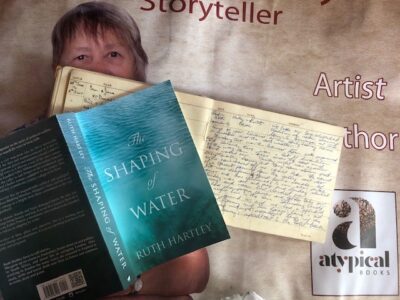 Photo of the book The Shaping of Water laid open across the cottage guest-book on which it was based. Both books lie over a poster of Ruth Hartely showing her "atypical books" imprint in the right hand bottom corner.